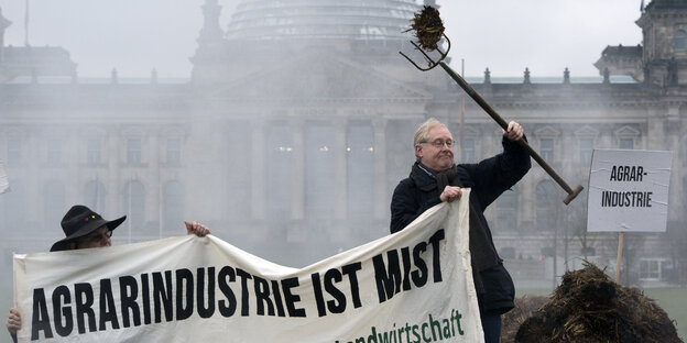 Ein Mann mit einer Mistgabel hält ein Transparent mit dem Aufdruck "Agrarindustrie ist Mist" Ein Mann mit einer Mistgabel hält ein Transparent mit dem Aufdruck "Agrarindustrie ist Mist"