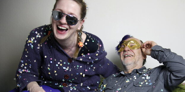junge Frau mit Sonnenbrille und ein älterer Mann mit Maske sitzen auf einem Sofa und lachen. Sie haben Konfetti auf ihrer Kleidung junge Frau mit Sonnenbrille und ein älterer Mann mit Maske sitzen auf einem Sofa und lachen. Sie haben Konfetti auf ihrer Kleidung
