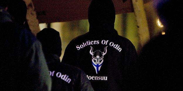 Männer stehen im Dunkeln in einer finnischen Stadt. Auf dem Rücken ihrer Jacken steht „Soldiers of Odin“ Männer stehen im Dunkeln in einer finnischen Stadt. Auf dem Rücken ihrer Jacken steht „Soldiers of Odin“