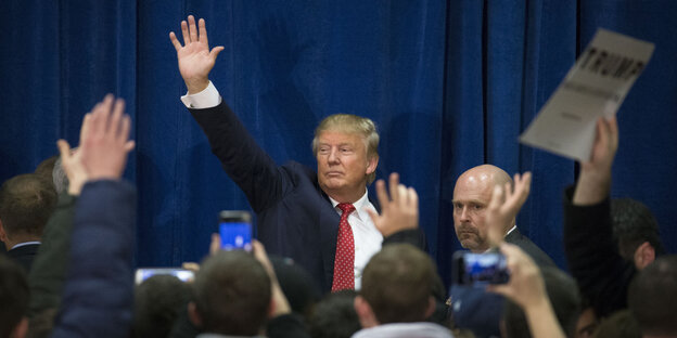 Mann im Anzug, es ist Donald Trump, hebt seine Hand, vor ihm lauter Menschen, teilweise auch mit in die Höhe gestreckten Armen Mann im Anzug, es ist Donald Trump, hebt seine Hand, vor ihm lauter Menschen, teilweise auch mit in die Höhe gestreckten Armen