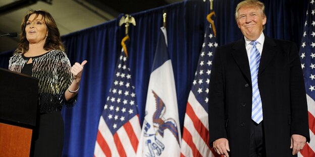 Eine Frau, Sarah Palin, steht auf einer Bühne und spricht, sie zeigt mit dem Daumen auf einen Mann, Donald Trumpl, der neben ihr stet Eine Frau, Sarah Palin, steht auf einer Bühne und spricht, sie zeigt mit dem Daumen auf einen Mann, Donald Trumpl, der neben ihr stet