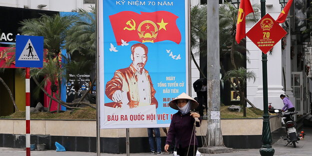 Ein Mensch mit Hut und Mundschutz läuft auf der Straße vor einem Plakat