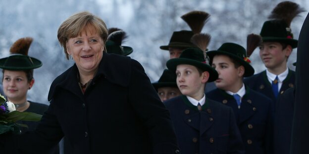 Die Kanzlerin steht draußen und schaut zur Seite. Hinter ihr stehen Kinder in bayrischen Trachten. Die Kanzlerin steht draußen und schaut zur Seite. Hinter ihr stehen Kinder in bayrischen Trachten.