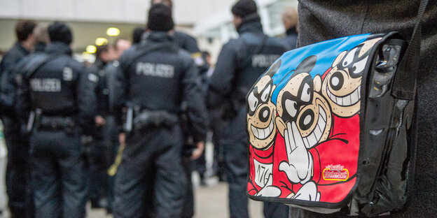 Im Hintergrund sieht man Polizisten. Im Vordegrund steht ein Mensch, der eine Umhängetasche mit Panzerknacker-Motiv trägt Im Hintergrund sieht man Polizisten. Im Vordegrund steht ein Mensch, der eine Umhängetasche mit Panzerknacker-Motiv trägt