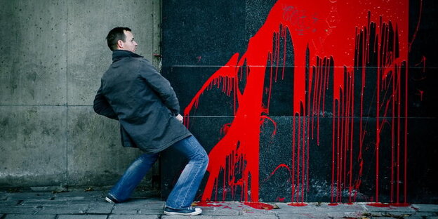 Ein Mann steht in Pinkelpose vor einer mit rotem Graffiti besprühten Wand Ein Mann steht in Pinkelpose vor einer mit rotem Graffiti besprühten Wand
