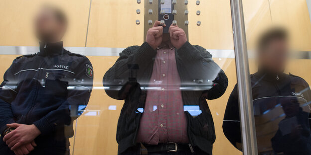 Der Angeklagte Nils D. mit einem Aktenordner vorm Gesicht hinter Sicherheitsglas Der Angeklagte Nils D. mit einem Aktenordner vorm Gesicht hinter Sicherheitsglas