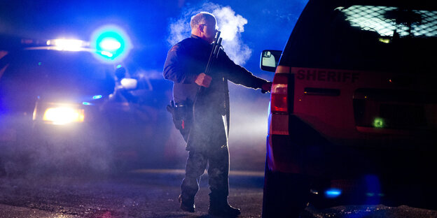 Ein amerikanischer Polizist trägt eine Waffe in ein Auto, es ist dunkel, hinter ihm Blaulicht. Ein amerikanischer Polizist trägt eine Waffe in ein Auto, es ist dunkel, hinter ihm Blaulicht.