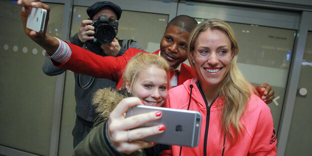 Eine strahlend lächelnde Frau mit blonden Haaren und pinker Sportjacke - es ist Angelique Kerber - vor ihr eine Frau mit Handy, hinter ihr ein Mann mit Handy, beide versuchen sich mit Kerber zu fotografieren Eine strahlend lächelnde Frau mit blonden Haaren und pinker Sportjacke - es ist Angelique Kerber - vor ihr eine Frau mit Handy, hinter ihr ein Mann mit Handy, beide versuchen sich mit Kerber zu fotografieren
