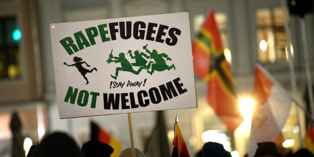 Pegida-Demonstranten im Hintergrund. Im Vordergrund ein Schild, auf dem „Rapefugees not Welcome“ steht. Pegida-Demonstranten im Hintergrund. Im Vordergrund ein Schild, auf dem „Rapefugees not Welcome“ steht.
