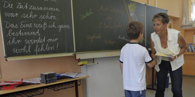 Grundschullehrerin gibt Schüler die Hand vor Tafel.