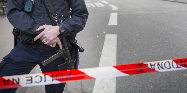 rot-weißes Absperrband, dahitner ein Polizist mit Maschinengewehr, sichtbar ab unterhalb der Schulter. rot-weißes Absperrband, dahitner ein Polizist mit Maschinengewehr, sichtbar ab unterhalb der Schulter.
