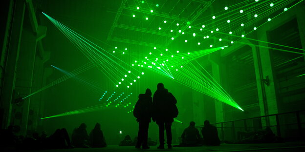 Menschen stehen und sitzen in einer großen Halle. Von der Decke strahlen viele Laser in den Raum. Menschen stehen und sitzen in einer großen Halle. Von der Decke strahlen viele Laser in den Raum.