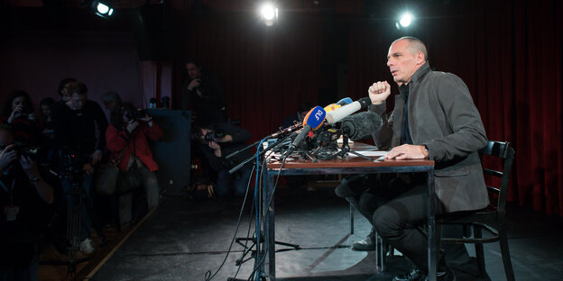 Yanis Varoufakis sitzt an einem Tisch vor einem Mikrofon, erleuchtet von Spots, sonst ist es dunkel Yanis Varoufakis sitzt an einem Tisch vor einem Mikrofon, erleuchtet von Spots, sonst ist es dunkel