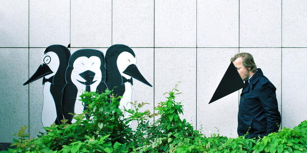 Etwas abseits von drei Pinguinen, die an eine Wand gemalt sind, steht ein Mann, der sich einen Schnabel aufgesetzt hat Etwas abseits von drei Pinguinen, die an eine Wand gemalt sind, steht ein Mann, der sich einen Schnabel aufgesetzt hat