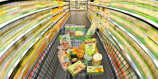 Ein Einkaufswagen mit Lebensmitteln wird durch einen Supermarkt geschoben (Aufnahme mit langer Verschlusszeit) Ein Einkaufswagen mit Lebensmitteln wird durch einen Supermarkt geschoben (Aufnahme mit langer Verschlusszeit)