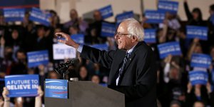 Bernie Sanders am Redpult, im Hintergrund viele Menschen mit blauen Schildern, er lächelt und hebt die Hand.