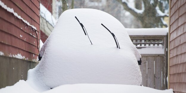 Ein Auto ist komplett von Schnee bedeckt. Es steht in einer Lücke zwischen zwei Häusern.