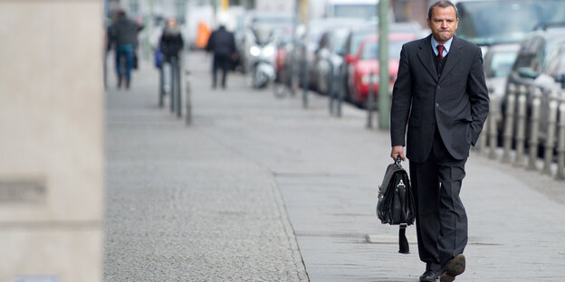 Edathy geht die Straße entlang, mit einem Koffer in der Hand und guckt grimmig.