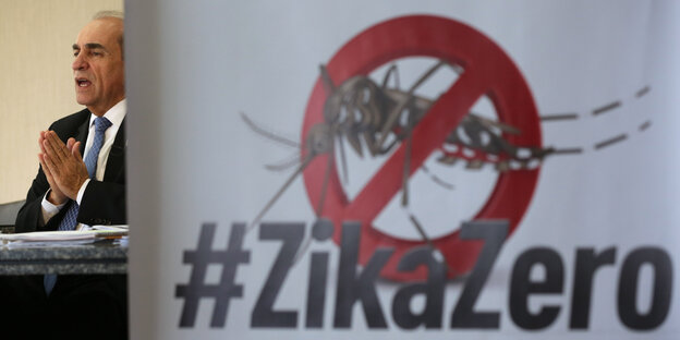 Ein Mann im Anzug hält seine Hände zusammengelegt hoch, neben ihm ein Schild, auf dem eine durchgestrichene Zika-Fliege und °'Zikozero" abgebildet ist Ein Mann im Anzug hält seine Hände zusammengelegt hoch, neben ihm ein Schild, auf dem eine durchgestrichene Zika-Fliege und °'Zikozero" abgebildet ist
