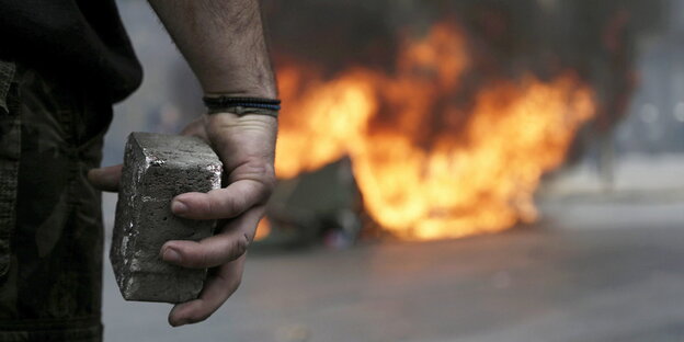 Im Vordergrund der Ausschnitt eines Armes und einer Hand, die einen Backstein hält, dahinter eine Straße, auf der ein Feuer brennt.