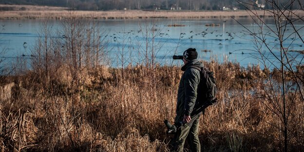 Ein Mann steht mit Fernglas in einer winterlichen Landschaft und beobachtet Vögel auf einem See.