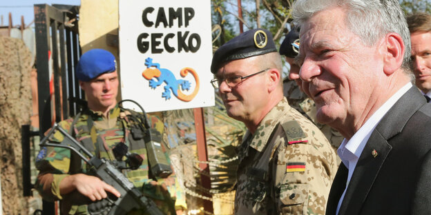 Herr Gauck steht neben zwei Bundeswehrsoldaten und dem Schild „Camp Gecko“. Herr Gauck steht neben zwei Bundeswehrsoldaten und dem Schild „Camp Gecko“.
