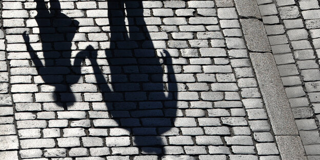 Der Schatten eines Erwachsenen, der die Hand eines Kindes hält, fällt auf Kopfsteinpflaster Der Schatten eines Erwachsenen, der die Hand eines Kindes hält, fällt auf Kopfsteinpflaster