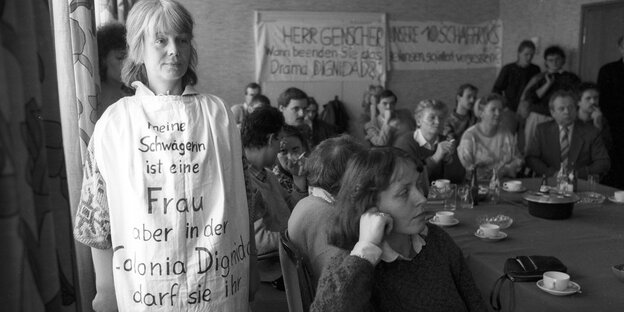 Schwarz-Weiß-Aufnahme einer Frau mit einem beschrifteten Leibchen, im Hintergrund sitzen viele Menschen an langen Tischen.