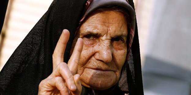 Eine alte Frau mit schwarzem Kopftuch hebt ihre Hand zum Victory-Zeichen. Eine alte Frau mit schwarzem Kopftuch hebt ihre Hand zum Victory-Zeichen.