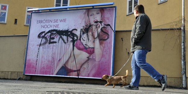 Auf einem mit „Sexismus“ beschmierten Plakat ist eine Frau in Dessous zu sehen. Ein Mann spaziert vorbei Auf einem mit „Sexismus“ beschmierten Plakat ist eine Frau in Dessous zu sehen. Ein Mann spaziert vorbei