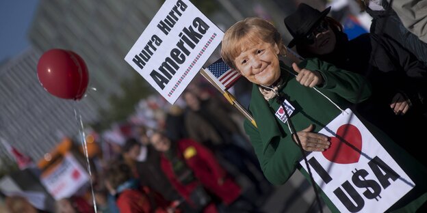 ein Demonstrant mit Merkel-Maske und zwei Schildern, auf einem steht „Hurra Hurra Amerika“, auf dem anderen „Ich liebe USA“ ein Demonstrant mit Merkel-Maske und zwei Schildern, auf einem steht „Hurra Hurra Amerika“, auf dem anderen „Ich liebe USA“