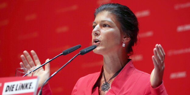 Sahra Wagenknecht im roten Kleid mit ausgebreiteten Armen vor rotem Hintergrund