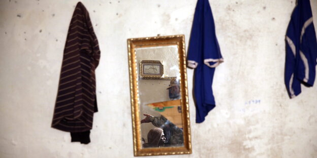 In einem Spiegel an einer Wand sieht man einen libyschen Soldaten und einen Flüchtling. Daneben hängen dunkle T-Shirts In einem Spiegel an einer Wand sieht man einen libyschen Soldaten und einen Flüchtling. Daneben hängen dunkle T-Shirts