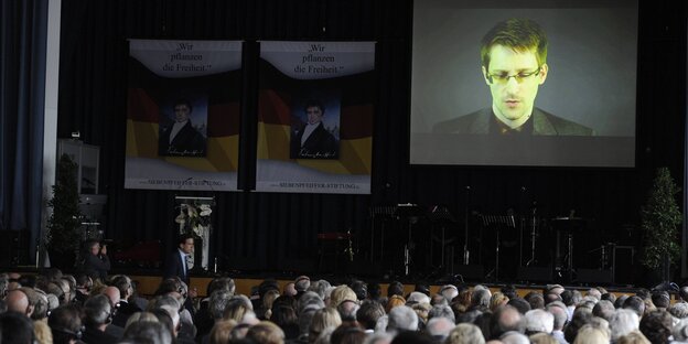 Menschen sitzen in einem Saal, an der Wand eine Leinwand mit dem projizierten Konterfei Edward Snowdens Menschen sitzen in einem Saal, an der Wand eine Leinwand mit dem projizierten Konterfei Edward Snowdens