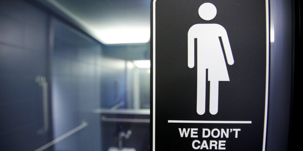 Ein Schild in einer Toilette zeigt die eine Person, deren Geschlecht nicht eindeutig ist und die Aufschrift „We don't care“ Ein Schild in einer Toilette zeigt die eine Person, deren Geschlecht nicht eindeutig ist und die Aufschrift „We don't care“