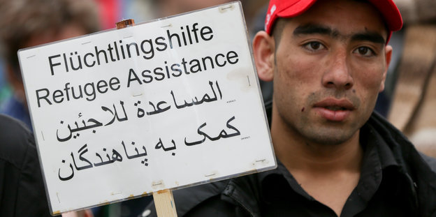 Ein Flüchtling hält ein Schild hoch auf dem in vier Sprachen "Flüchtlingshilfe" steht.
