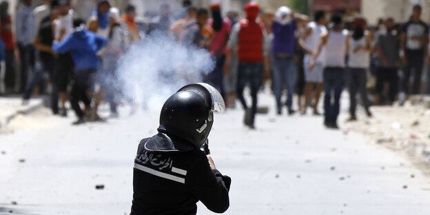 Ein Polizist mit Helm und arabischer Schrift auf der Jacke ist von hinten zu sehen, er feuert in Richtung einer Menschenmenge Ein Polizist mit Helm und arabischer Schrift auf der Jacke ist von hinten zu sehen, er feuert in Richtung einer Menschenmenge