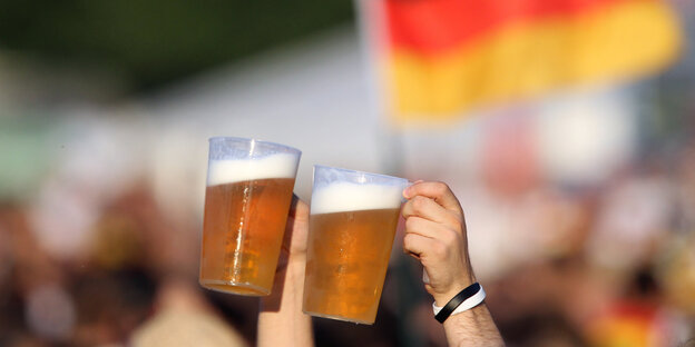 Zwei Bierbecher werden hochgehalten, im Hintergrund eine Menschenmenge und eine Deutschlandfahne Zwei Bierbecher werden hochgehalten, im Hintergrund eine Menschenmenge und eine Deutschlandfahne