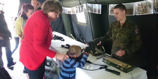 ein Kind mit einer Frau an einem Stand der Bundeswehr, das Kind hält ein Gewehr in der Hand, hinter dem Stand ein Mann in Uniform ein Kind mit einer Frau an einem Stand der Bundeswehr, das Kind hält ein Gewehr in der Hand, hinter dem Stand ein Mann in Uniform
