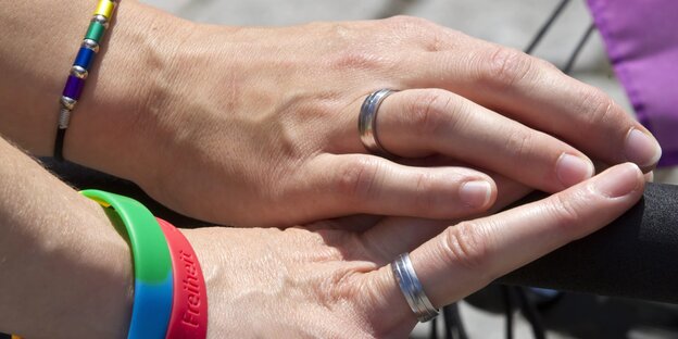 Zwei Hände mit Eheringen. An den Armen Regenbogen-Armbänder Zwei Hände mit Eheringen. An den Armen Regenbogen-Armbänder