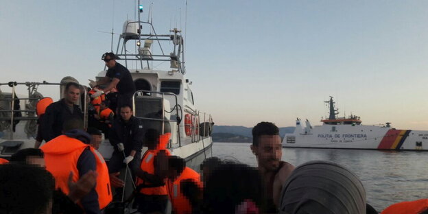 Flüchtlinge mit Mitgliedern der Küstenwache in orangefarbenen Warnwesten auf einem Boot, im Hintergrund das Frontex-Boot Flüchtlinge mit Mitgliedern der Küstenwache in orangefarbenen Warnwesten auf einem Boot, im Hintergrund das Frontex-Boot