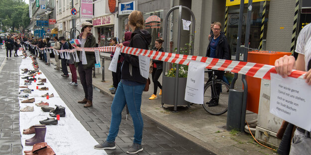 Menschen stehen entlang einem rot-weißen Absperrband, davor eine weißer Streifen Papier, auf dem Schuhe aufgereiht sind Menschen stehen entlang einem rot-weißen Absperrband, davor eine weißer Streifen Papier, auf dem Schuhe aufgereiht sind