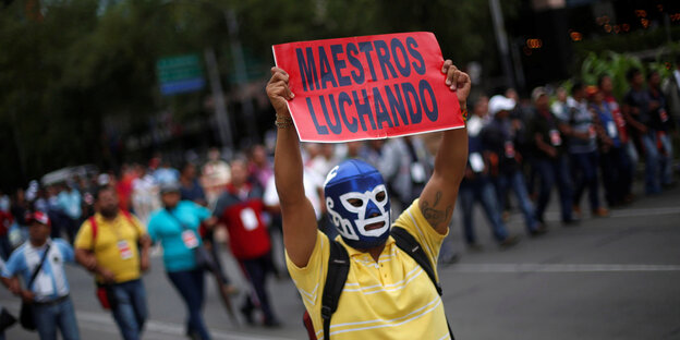 Ein Demonstrant trägt eine Wrestlingmaske des Lucha Libre und hält ein Schild hoch, auf dem „Maestros Luchando“ steht Ein Demonstrant trägt eine Wrestlingmaske des Lucha Libre und hält ein Schild hoch, auf dem „Maestros Luchando“ steht