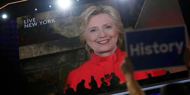 Hillary Clinton in einer Videoübertragung, im Vordergrund hält jemand ein Schild, auf dem „History“ steht Hillary Clinton in einer Videoübertragung, im Vordergrund hält jemand ein Schild, auf dem „History“ steht
