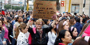 Inmitten einer vorwärts gehenden Menschenmenge hält eine Frau mit kurzen Haaren und Brille ein Schild hoch.