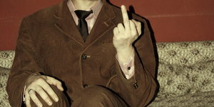 Ein Mann im braunen Anzug sitzt auf einem Sofa und zeigt den Mittelfinger