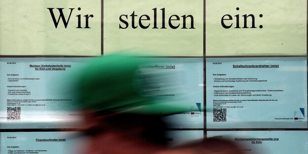 Stellenanzeigen mit der Überschrift "Wir stellen ein" hängen im Fenster einer Zeitarbeitsfirma und eine Person läuft vorbei Stellenanzeigen mit der Überschrift "Wir stellen ein" hängen im Fenster einer Zeitarbeitsfirma und eine Person läuft vorbei