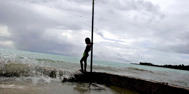 Ein Mensch lehnt an einem Pfahl, eine Welle Meerwasser schwammt daneben über den Strand Ein Mensch lehnt an einem Pfahl, eine Welle Meerwasser schwammt daneben über den Strand