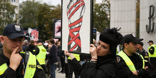 Ein in schwarz gekleidete Frau hält ein Bild einer Frau mit roten Fesseln hoch, um sie herum Polizisten in gelben Warnwesten