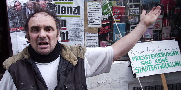 Ein älterer Mann mit Halbglate sitzt vor einem plakatierten Hintergrund und hält einen Arm hoch Ein älterer Mann mit Halbglate sitzt vor einem plakatierten Hintergrund und hält einen Arm hoch